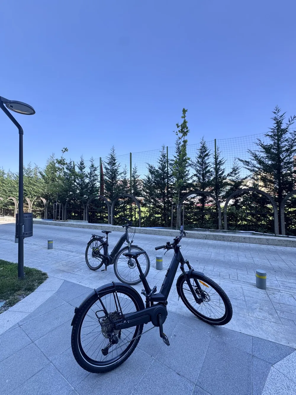 A black bicycle parked on a paved path against a bright blue sky, next to green trees and a building