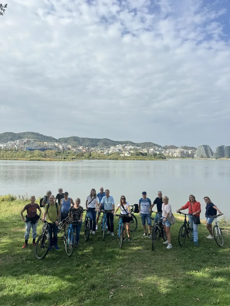 A large group of cyclists poses with their bikes on a grassy lakeshore with hills in the background