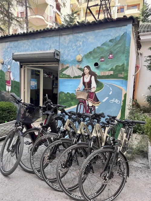 En række cykler parkeret foran butikken, udsmykket med et vægmaleri af en person i traditionelt albansk tøj