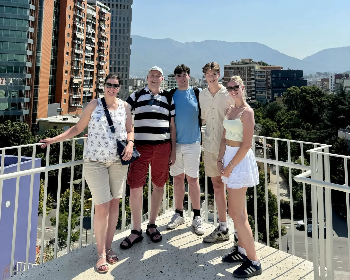 A group of five friends stands together on an elevated walkway with a scenic mountain range in the background