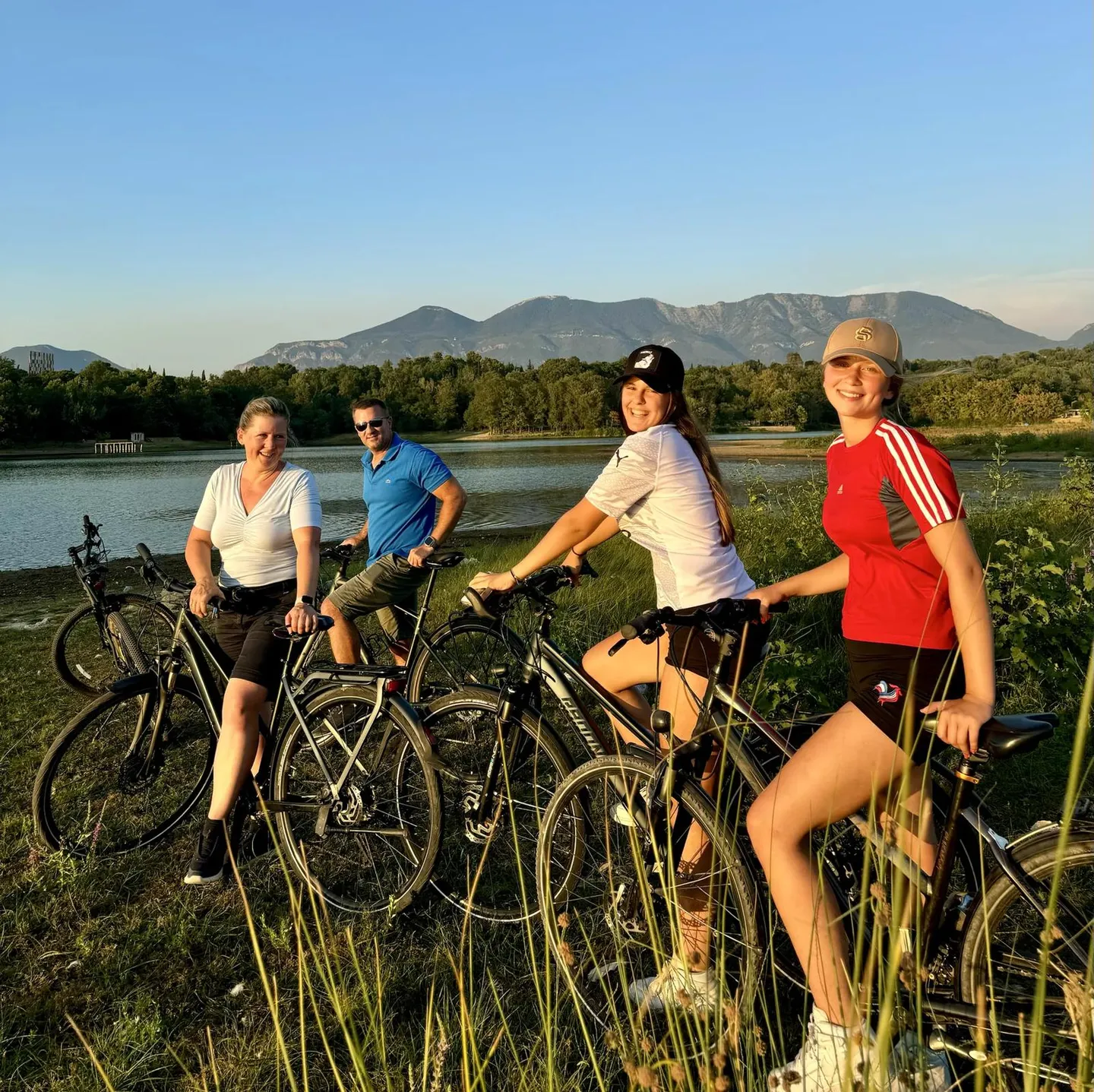 Four cyclists pose with their bikes on a grassy bank with hills and mountains in the background