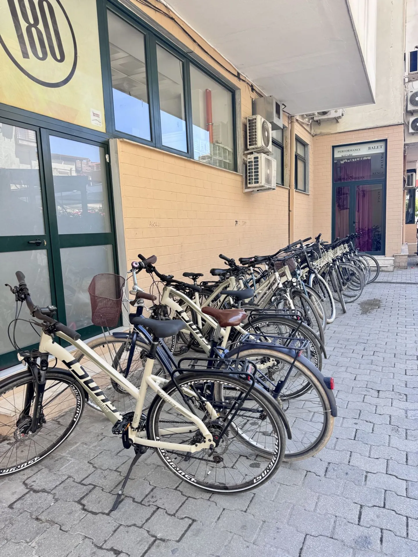 A row of beige rental bicycles parked on a tiled sidewalk next to the BikeTirana building